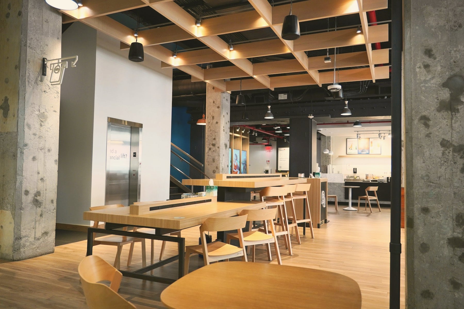 Espace commun de bureau : tables et chaises en bois, plafond à caissons, poteaux en béton