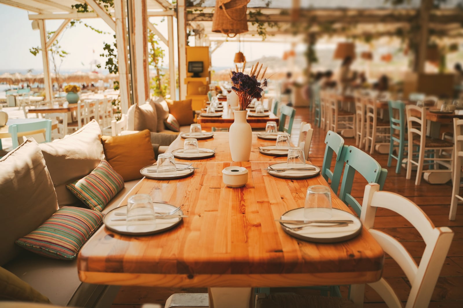 Terrasse de restaurant couverte : longue table en bois dressée, banquette à coussins et chaises en bois peint