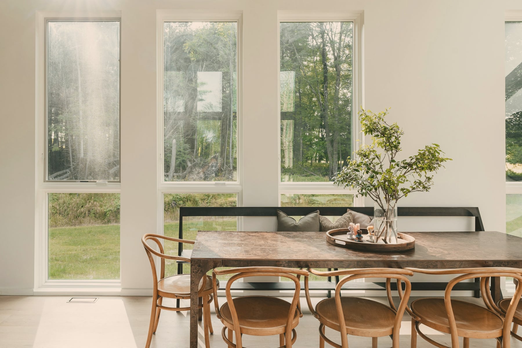 Salle à manger : table en bois sombre, banquette et chaises en bois courbé, grandes fenêtres sur la forêt