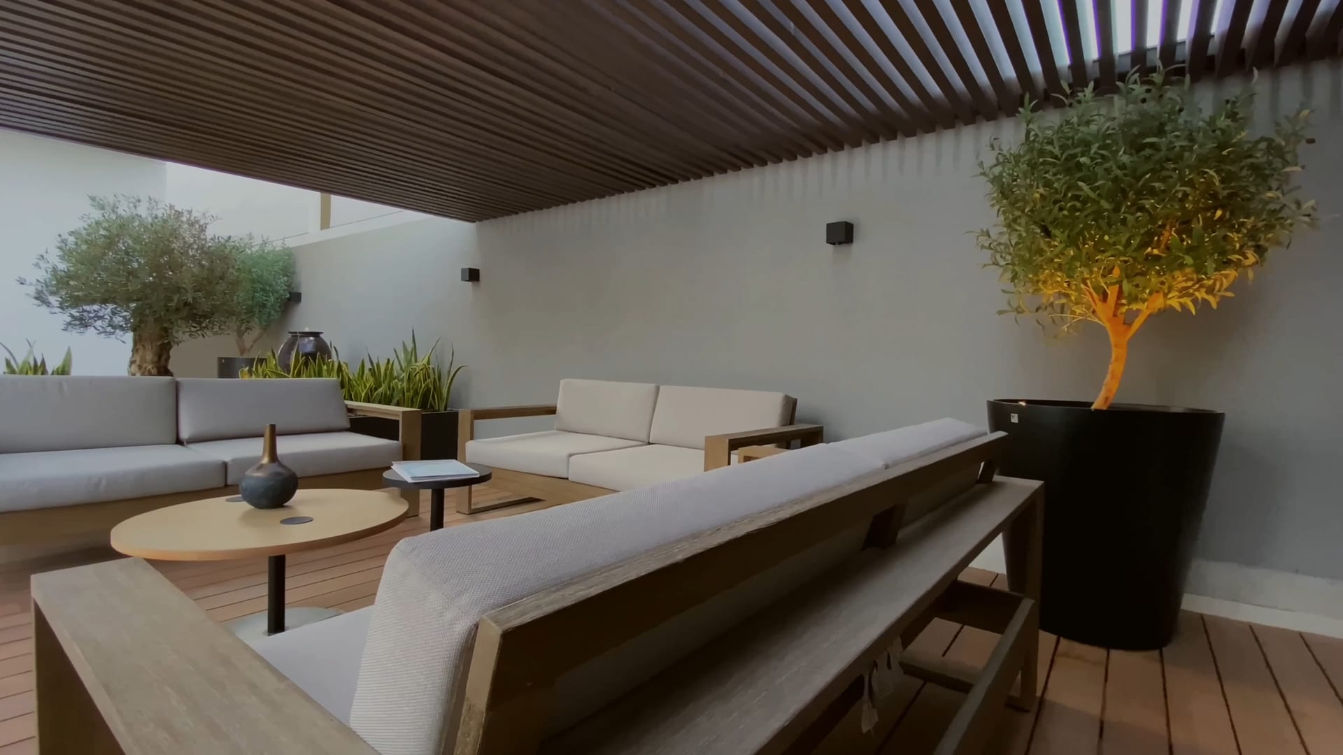 Salon d&rsquo;hôtel sous une pergola en bois, canapés clairs et olivier en pot, en fin de journée